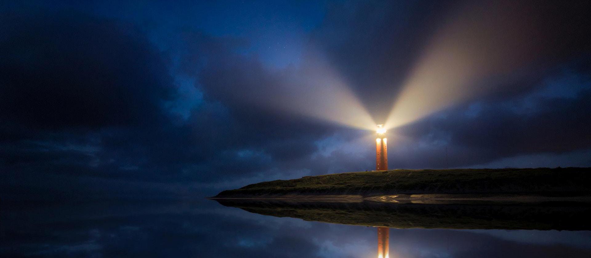 Image of lighted brown lighthouse beside body of water shining light out over the sea.